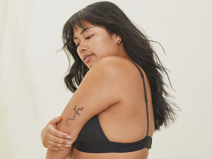 Person with long black hair wearing a black bra poses with their side facing the camera holding their arm. There is a tattoo on their arm.